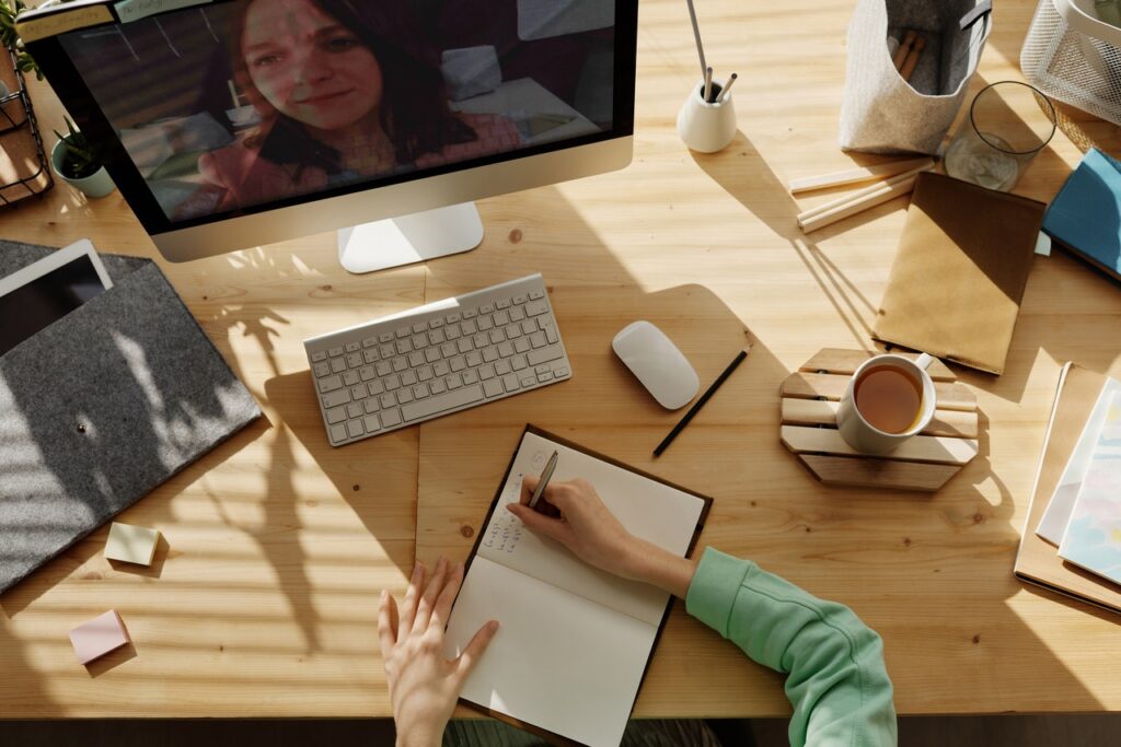 pessoa escrevendo no caderno e participando de uma reunião no computador